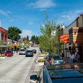 A small town street with parked cars on both sides. People walk along the sidewalk, past shops and restaurants. The sky is clear with a few clouds, and trees line parts of the street.