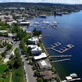 Aerial view of a lakeshore area with docks extending into the water. Boats are moored at the docks. Surrounding the lake are residential buildings, trees, and roads. The backdrop features a sprawling urban landscape under a partly cloudy sky.
