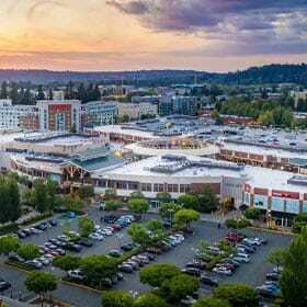 Aerial view of a large shopping center with several buildings and a parking lot filled with cars. Trees and green areas are scattered throughout. In the background, there are hills, residential buildings, and a colorful sunset sky.