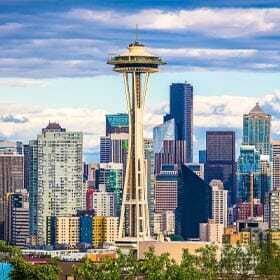 View of Seattle skyline featuring the Space Needle in the foreground, with various skyscrapers and buildings under a partly cloudy sky. Verdant trees are visible at the bottom of the image.