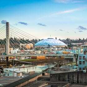 A cityscape featuring a large, white dome-shaped building, a tall cable-stayed bridge, and an assortment of buildings. The sky is clear with a few clouds, casting reflections on the water below.