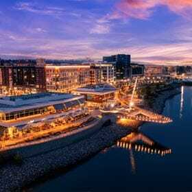 A vibrant waterfront cityscape at dusk, with illuminated buildings reflecting on the calm water. The sky is painted in shades of purple, pink, and orange. A lit pathway follows the curve of the shoreline, adding warmth to the scene.
