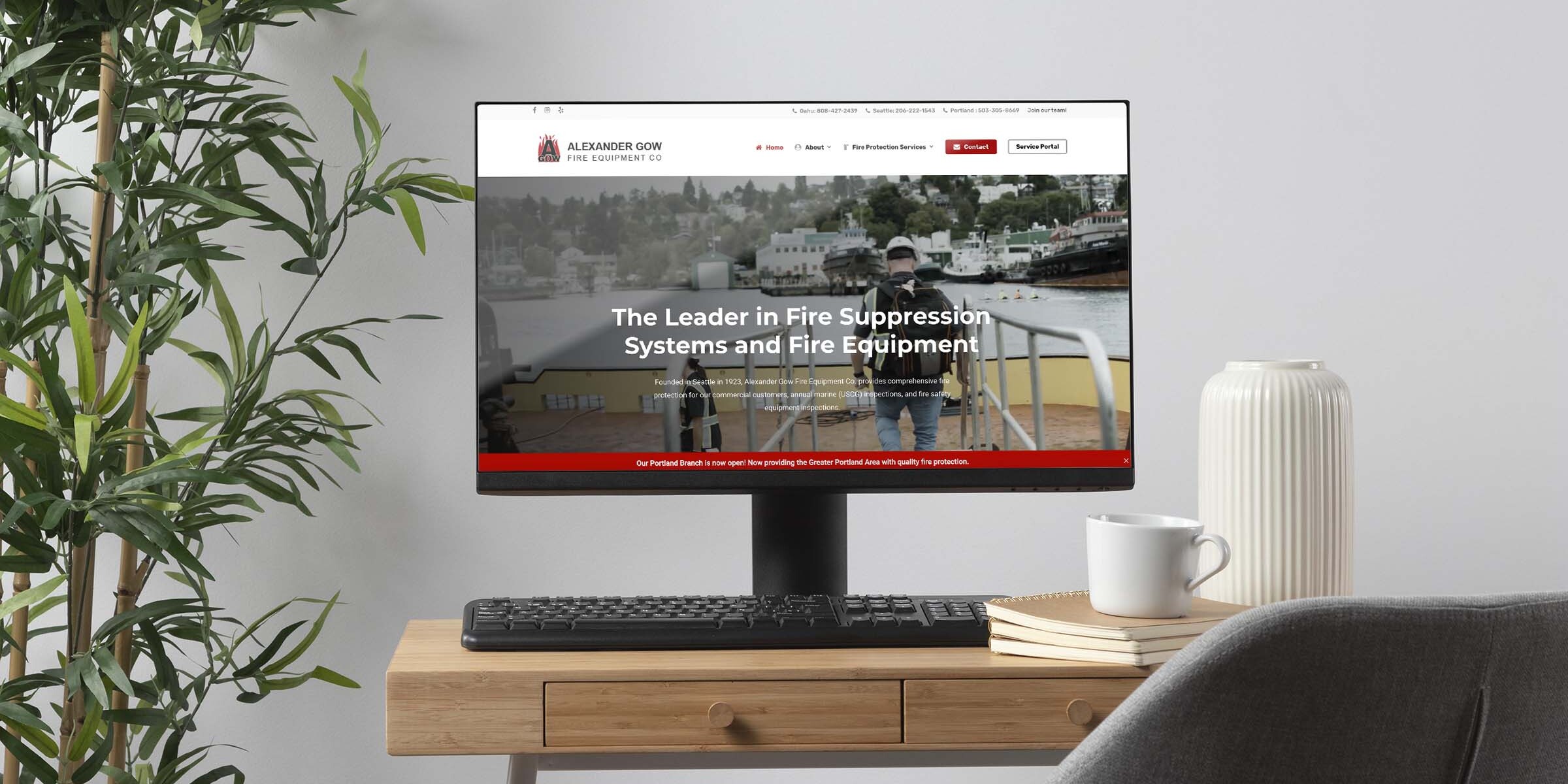 A computer on a wooden desk displays a website about fire suppression systems and equipment. A chair is beside the desk, with a white coffee mug and a white vase placed on the desk. A plant is partially visible on the left.