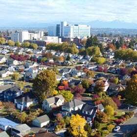 Aerial view of a suburban neighborhood with colorful autumn foliage. Houses are scattered among trees in various shades of red, yellow, and orange. In the background, a few tall buildings and distant mountains are visible under a cloudy sky.