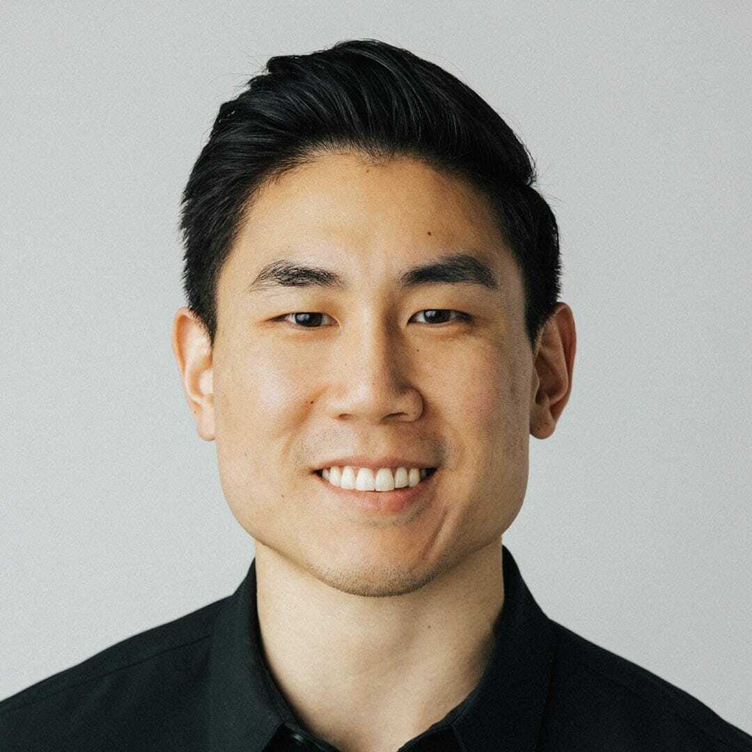 Portrait of a person smiling at the camera, wearing a black shirt. The background is plain and light-colored.
