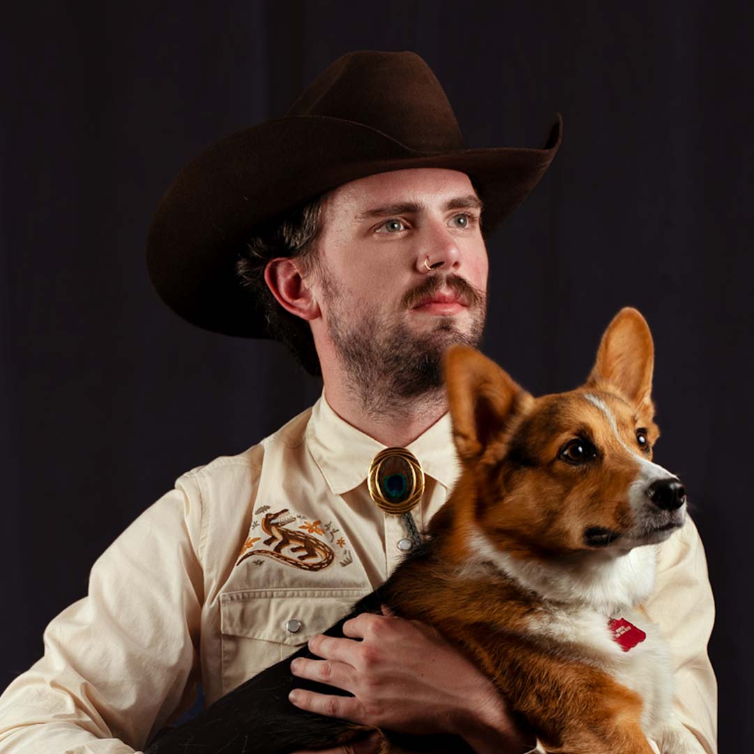 A person wearing a cowboy hat and embroidered shirt sits against a dark background, holding a Corgi dog. The person gazes forward, while the dog looks to the side.