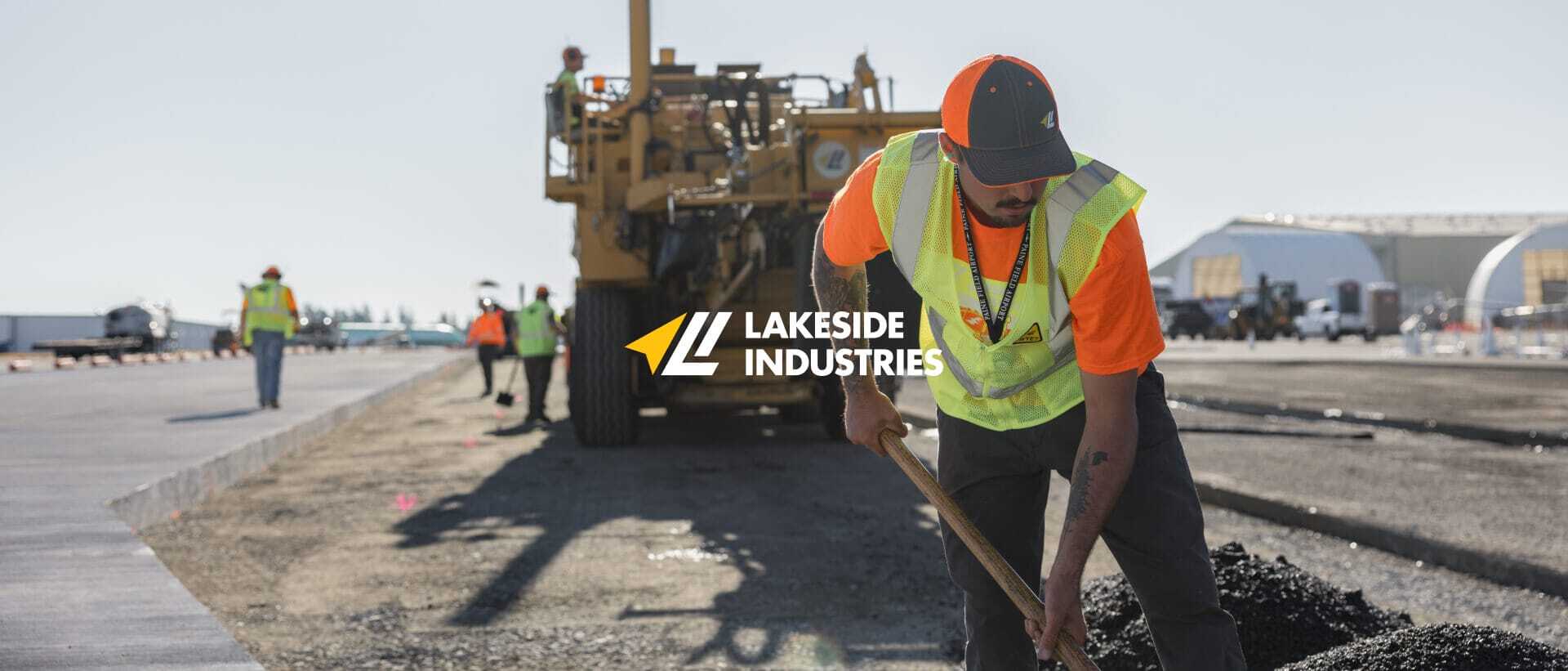 Worker in a neon vest and orange shirt uses a tool to smooth asphalt on a road. Large machinery and other workers are in the background. Lakeside Industries logo is overlaid on the image.