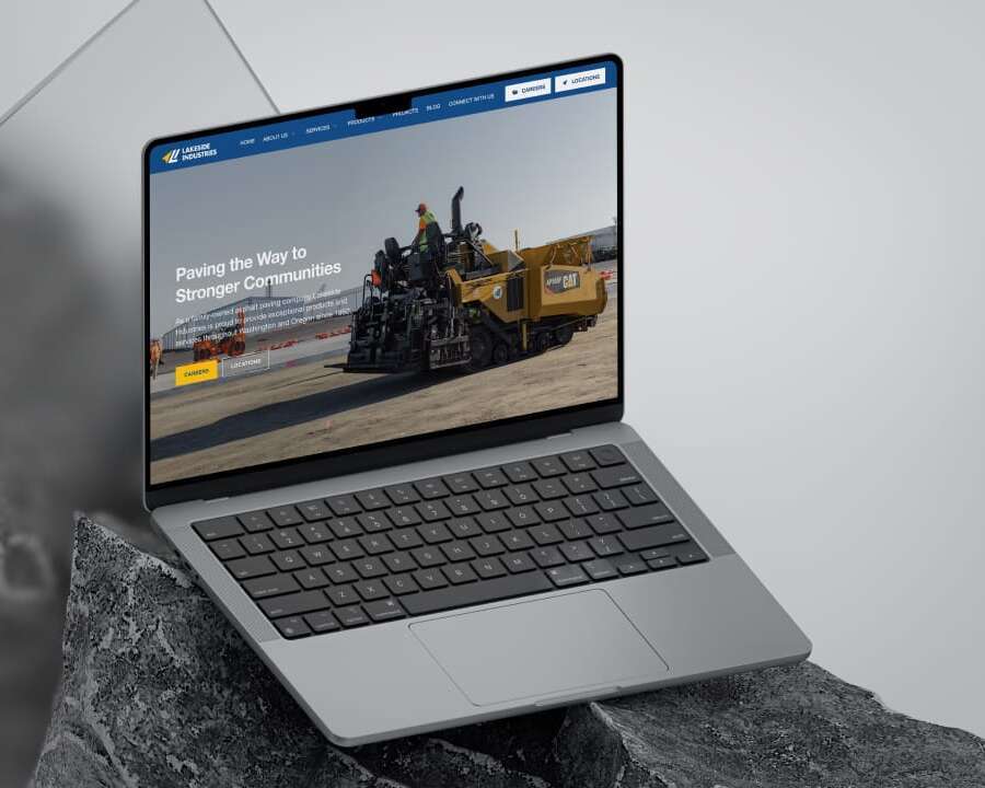 A laptop displaying a construction companys website, featuring an image of an asphalt paver on a road. The website tagline reads, Paving the Way to Stronger Communities. The laptop is positioned on a gray, textured surface.