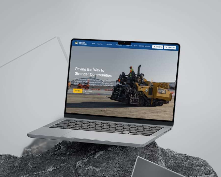A laptop displaying a website with an image of construction workers operating machinery on a road. The header reads, Paving the Way to Stronger Communities. The laptop is placed on a rocky surface with a sleek, modern design.