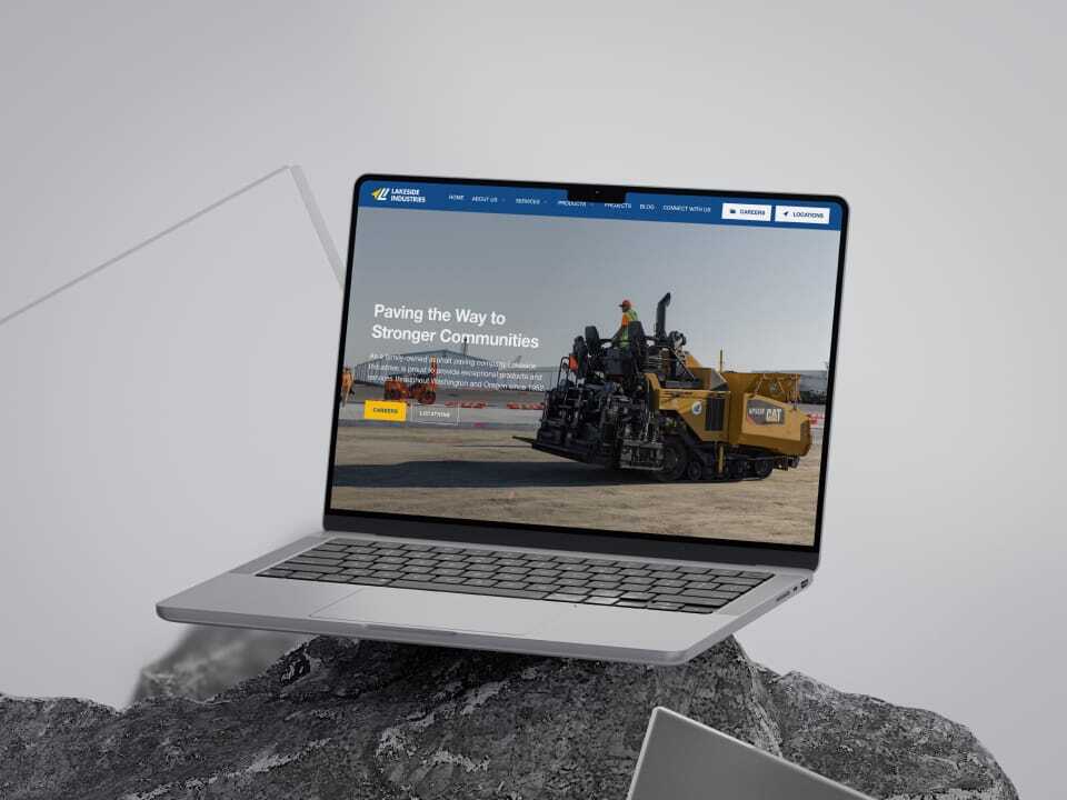 A laptop displaying a website with an image of construction workers operating machinery on a road. The header reads, Paving the Way to Stronger Communities. The laptop is placed on a rocky surface with a sleek, modern design.