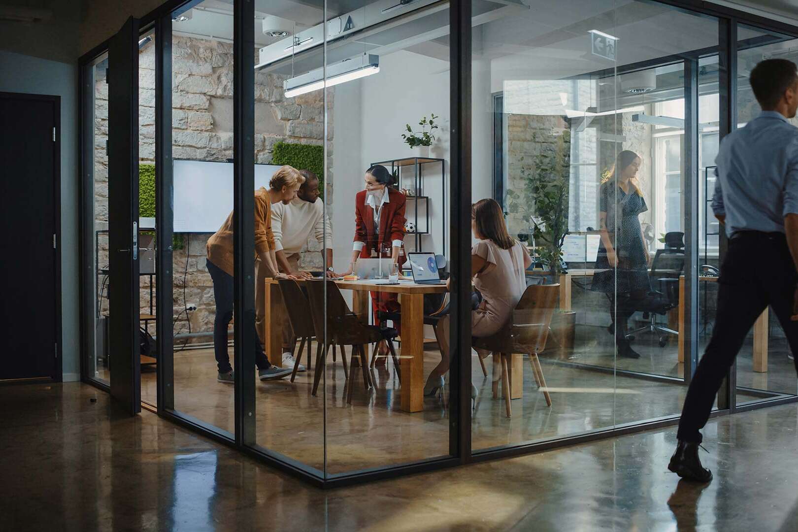 A group of people collaborate in a modern glass-walled meeting room with a wooden table and chairs. The space has stone walls and floor-to-ceiling windows. Outside, a person walks past, and another works at a desk.