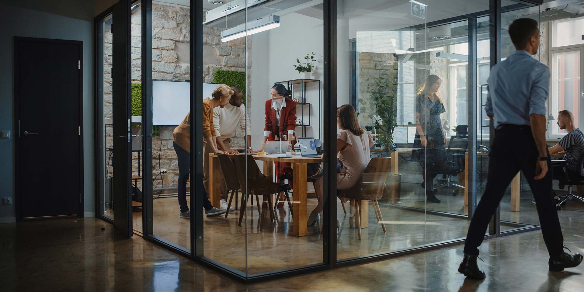 A group of people collaborate in a modern glass-walled meeting room with a wooden table and chairs. The space has stone walls and floor-to-ceiling windows. Outside, a person walks past, and another works at a desk.