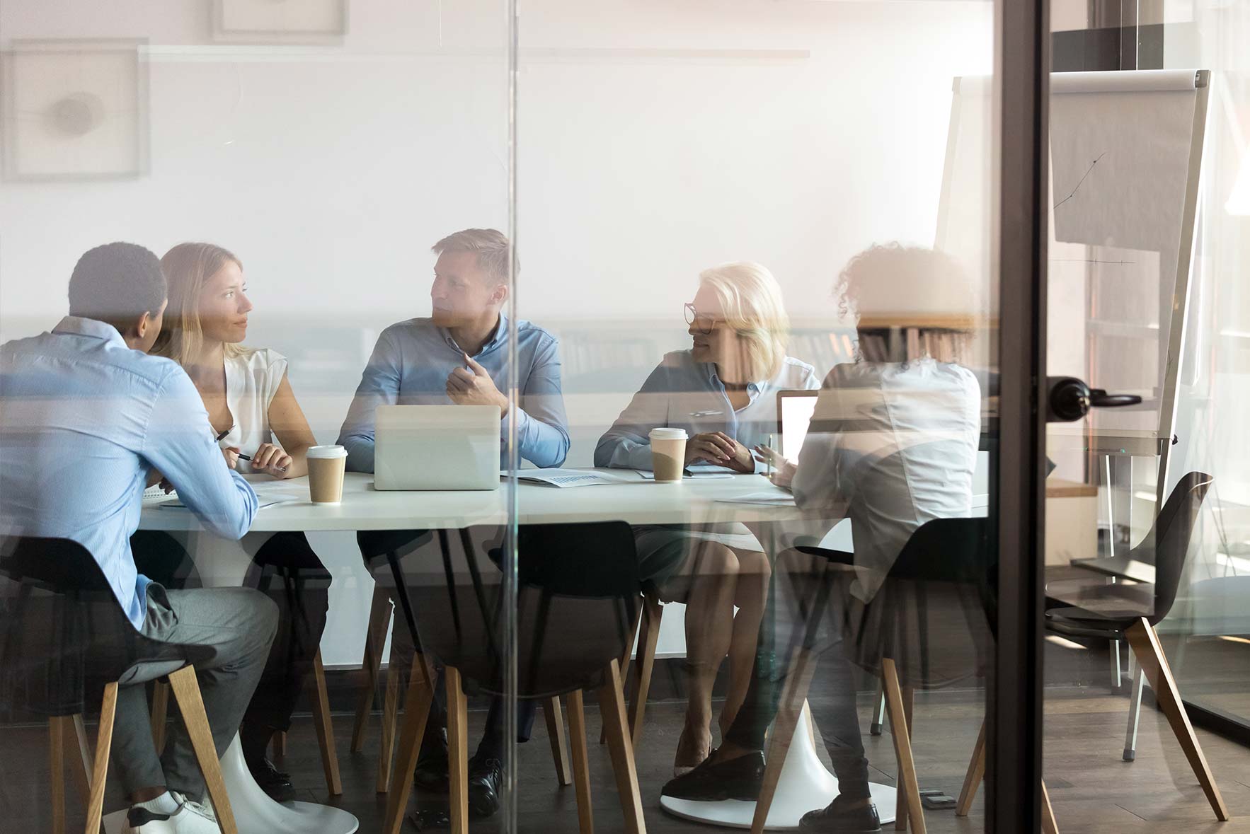A group of five people sit around a conference table in an office setting, engaged in discussion. Laptops and coffee cups are on the table. They are seen through a glass wall, with a flip chart in the background.