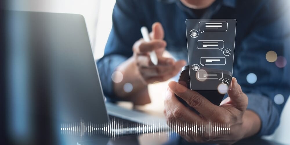 Person holding a smartphone with a digital chat interface displayed, seated in front of a laptop. They are smiling and holding a pen, suggesting a positive interaction. A sound wave graphic is visible at the bottom, implying audio elements.