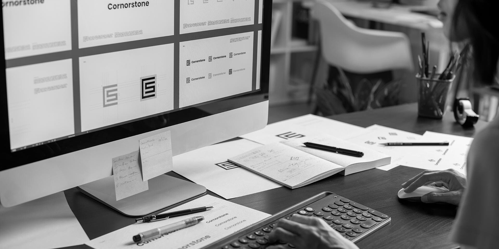 A person works at a desk, designing logos labeled Cornerstone on a computer screen. The desk has papers, pens, a keyboard, and a mouse. The workspace appears organized and focused on graphic design projects.