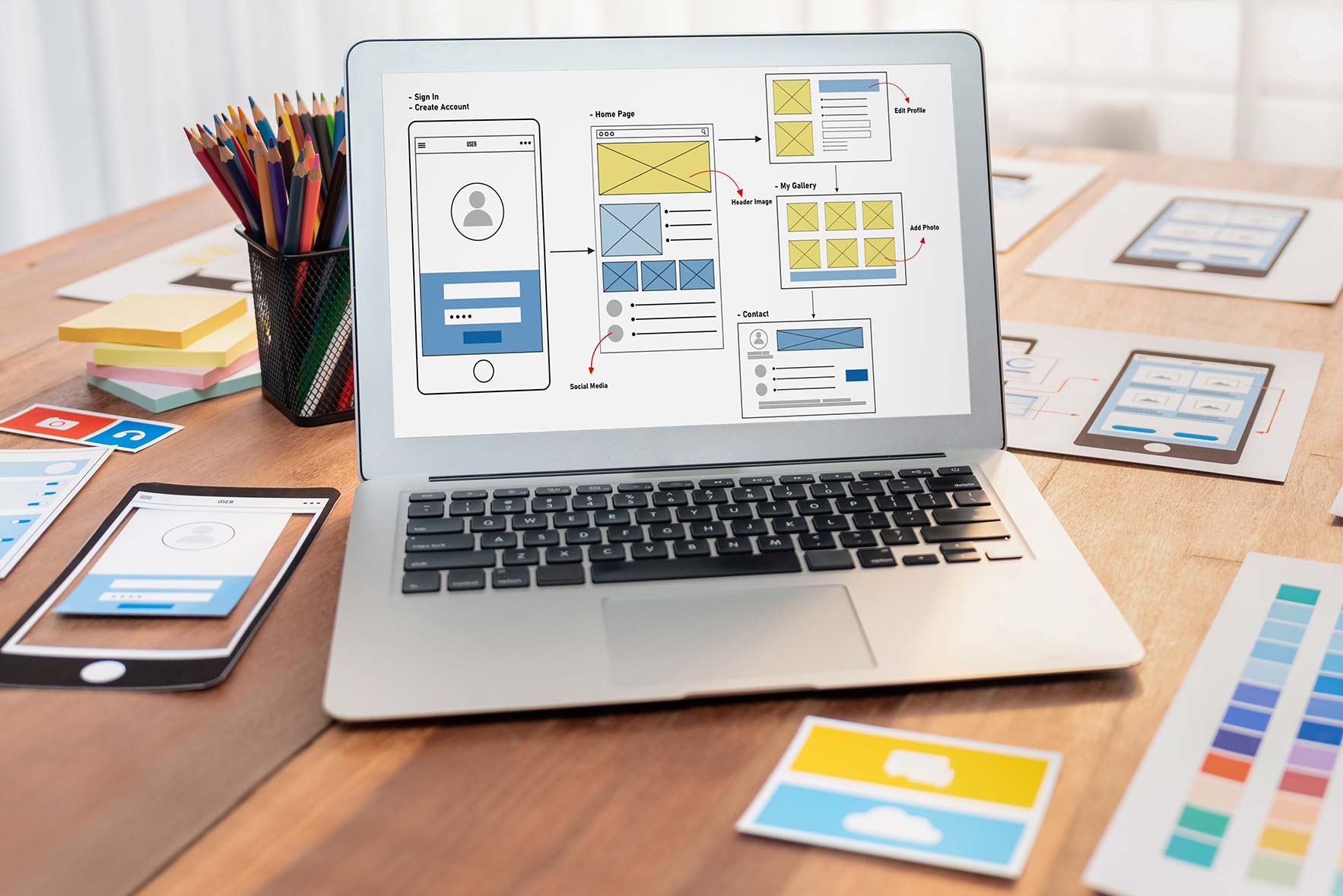 A laptop displays a wireframe design for a mobile app interface. The desk is cluttered with papers, colored pencils, and a smartphone with a similar design. Bright, natural light illuminates the workspace.