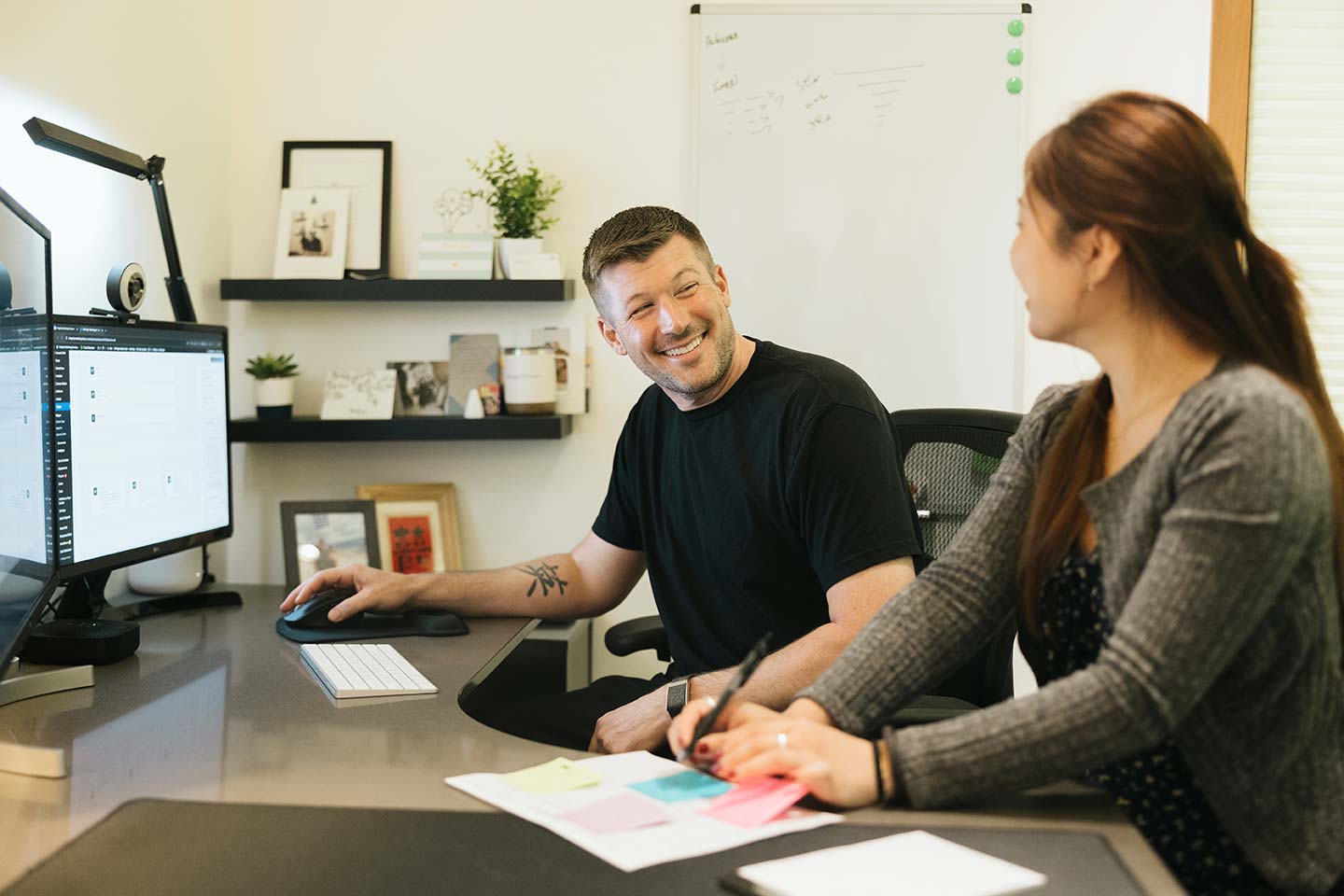 A man and woman are sitting at a desk in an office, smiling and talking. The man is using a computer with dual monitors. Papers with notes and a pen are on the desk. Shelves with decor and a whiteboard with writing are in the background.