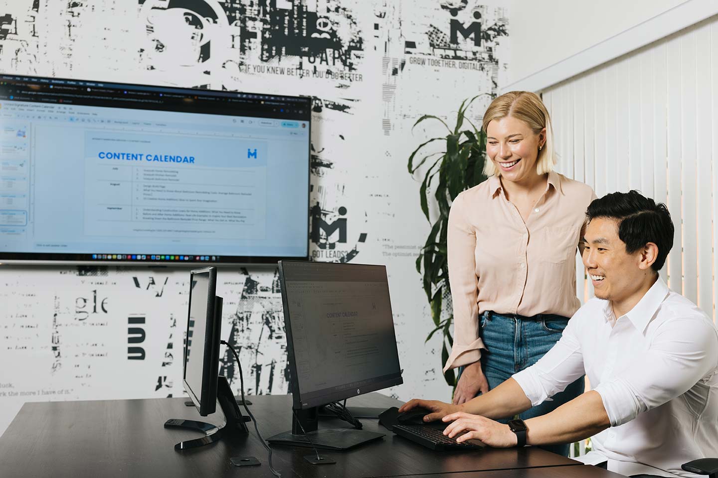 Two people are in an office setting. One is seated, working on a computer, smiling, while the other stands beside, looking at the screen. A large monitor on the wall displays a content calendar. The room has modern decor.