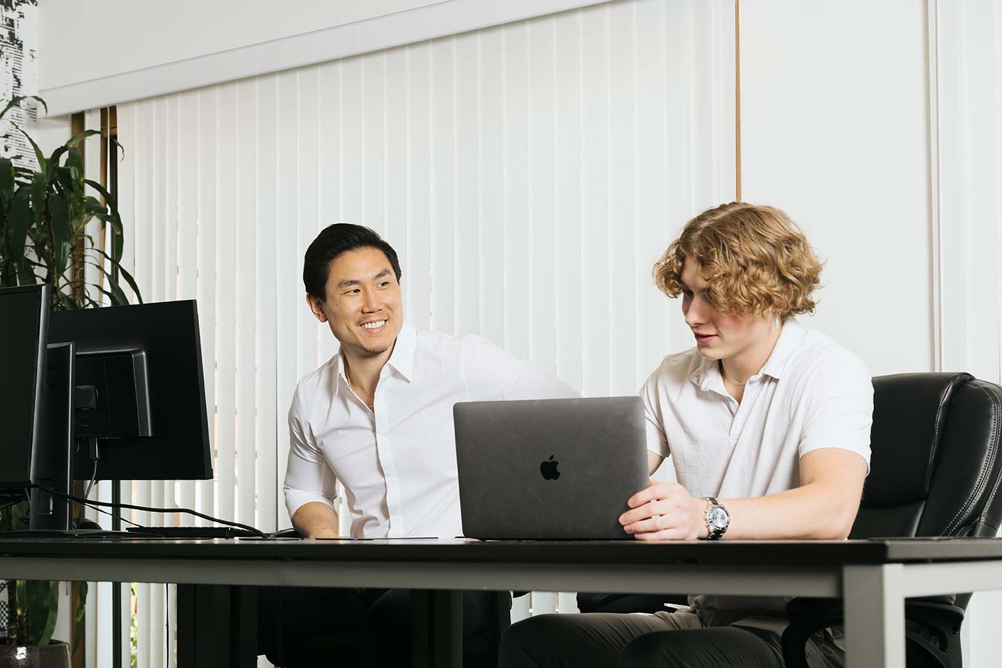 Two men in a bright office space sit beside each other. One man with dark hair smiles while the other with curly blonde hair focuses on a laptop. They sit at a desk with two monitors in front. Blinds cover the window in the background.