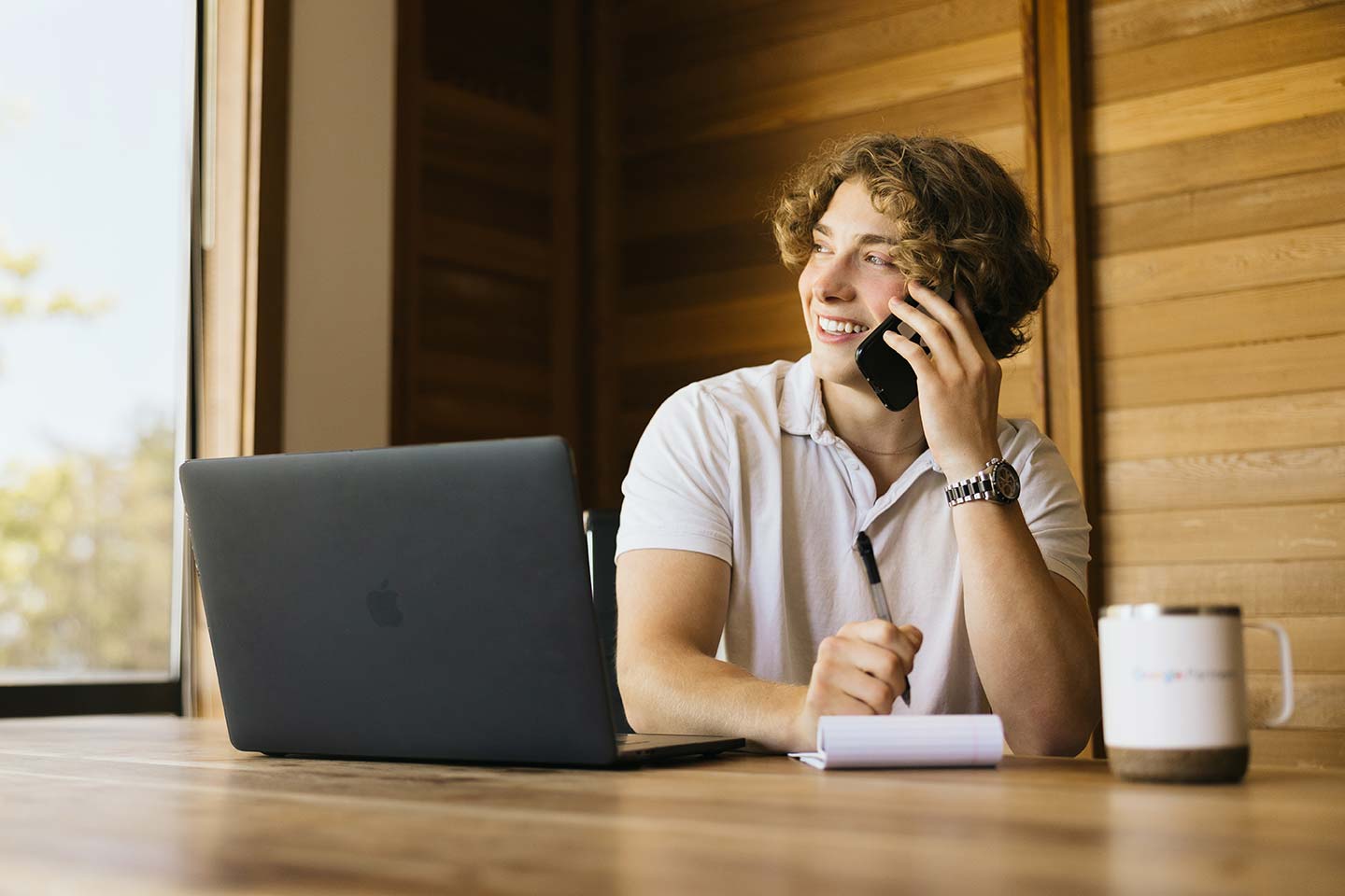 A person with curly hair is sitting at a table, smiling while talking on a phone. They have a laptop open in front of them and hold a pen with a notepad nearby. A coffee mug is on the table. Wood paneling is in the background.