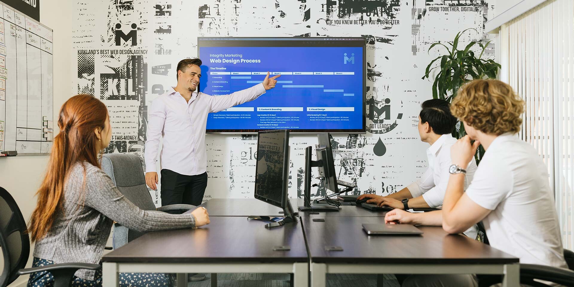A man is presenting in a conference room, pointing to a screen displaying a web design process. Three colleagues, one woman and two men, sit at a table with laptops, listening attentively. The room has a modern design with a patterned wall.