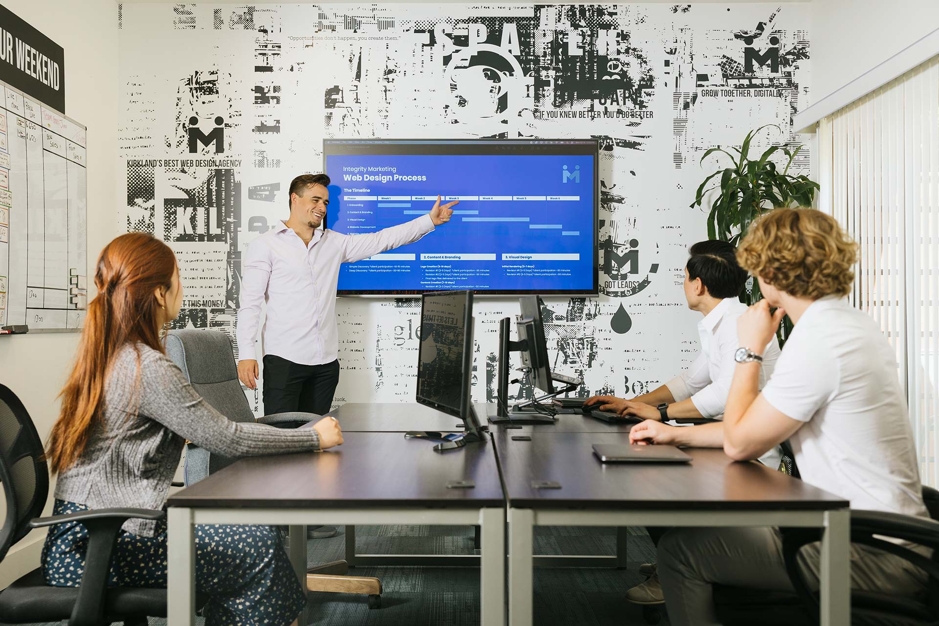A man is presenting in a conference room, pointing to a screen displaying a web design process. Three colleagues, one woman and two men, sit at a table with laptops, listening attentively. The room has a modern design with a patterned wall.