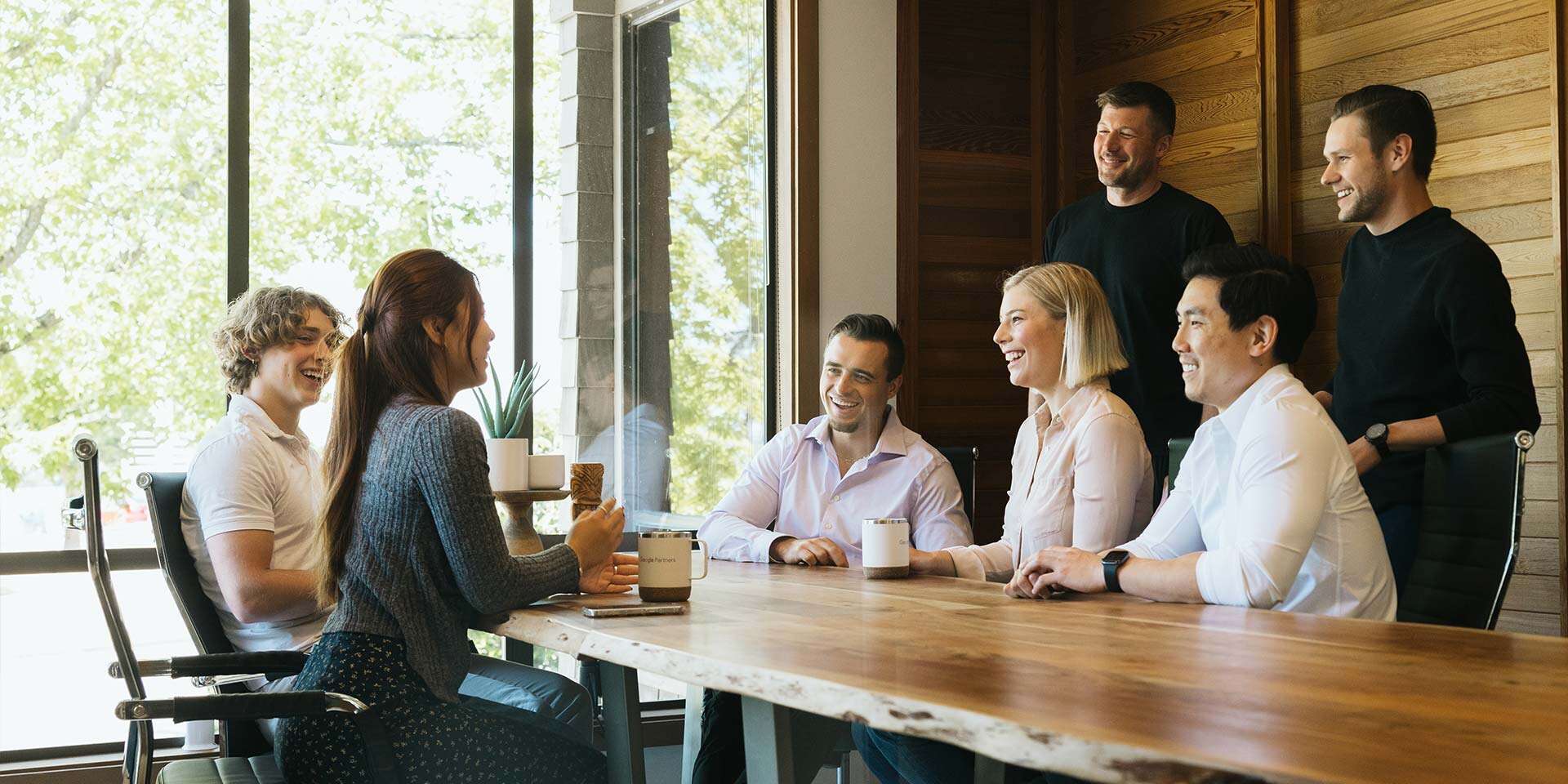 A group of six people sit and stand around a wooden table in a bright meeting room. They are engaged in conversation, smiling and holding mugs. Large windows with a view of trees are in the background.