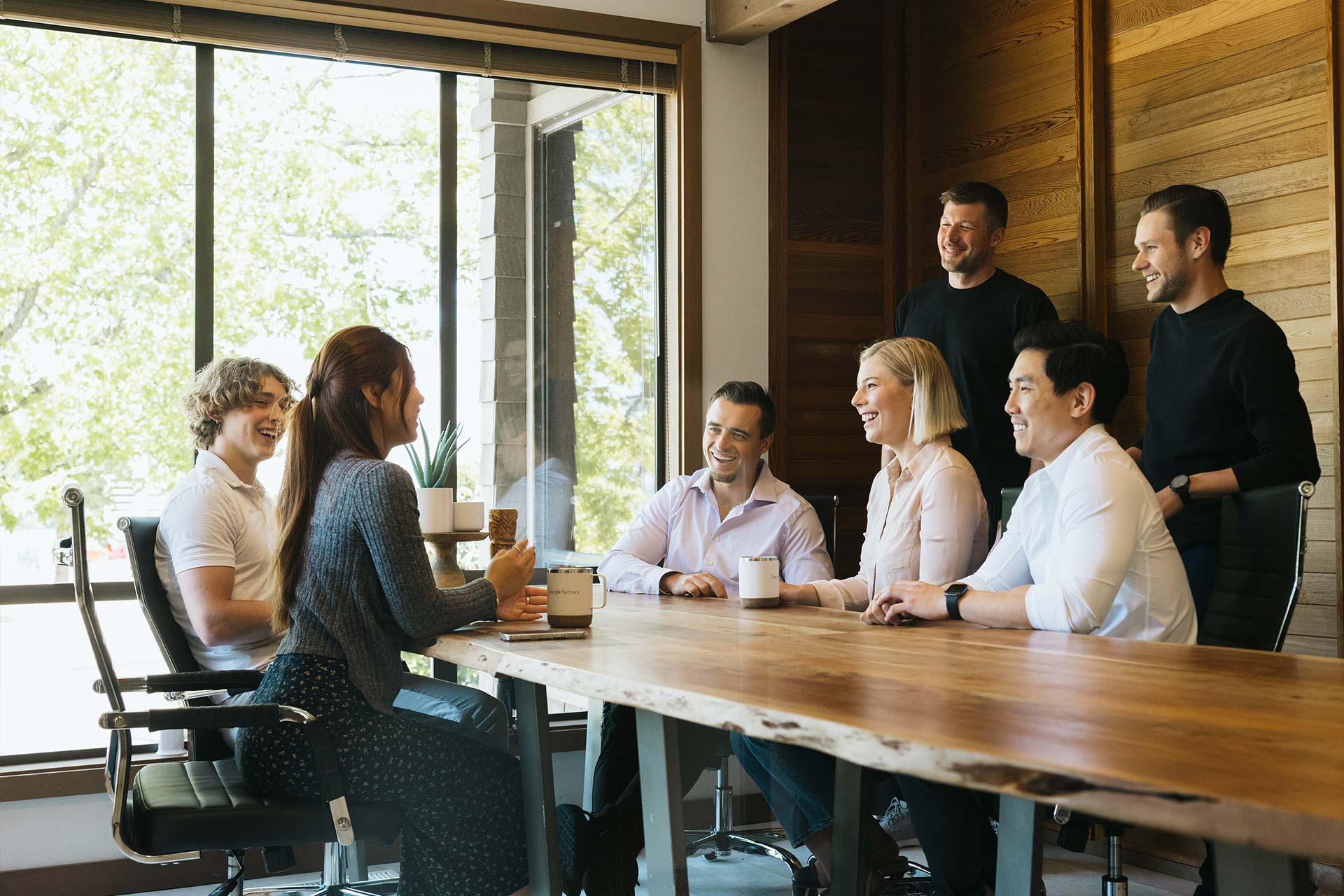 A group of six people sit and stand around a wooden table in a bright meeting room. They are engaged in conversation, smiling and holding mugs. Large windows with a view of trees are in the background.