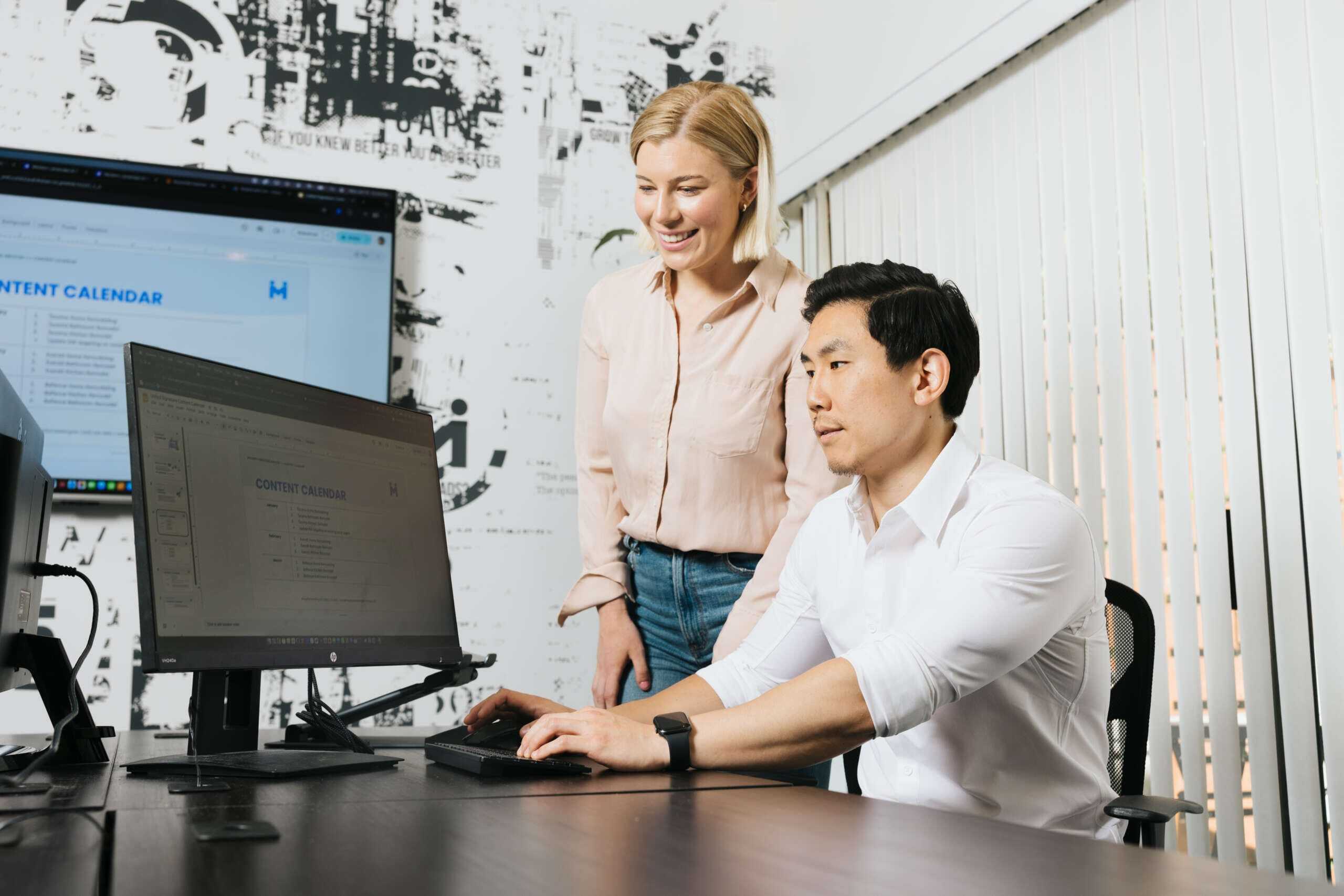 A man sits at an office desk, focused on a computer screen displaying a content calendar. A woman stands beside him, smiling and looking at the screen. The room has a modern design with a graphic wall and vertical blinds.