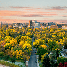 Aerial view of a city skyline with tall buildings at sunset. The foreground features a vast area of trees in vibrant autumn colors, including yellow, orange, and green, creating a serene and colorful landscape.