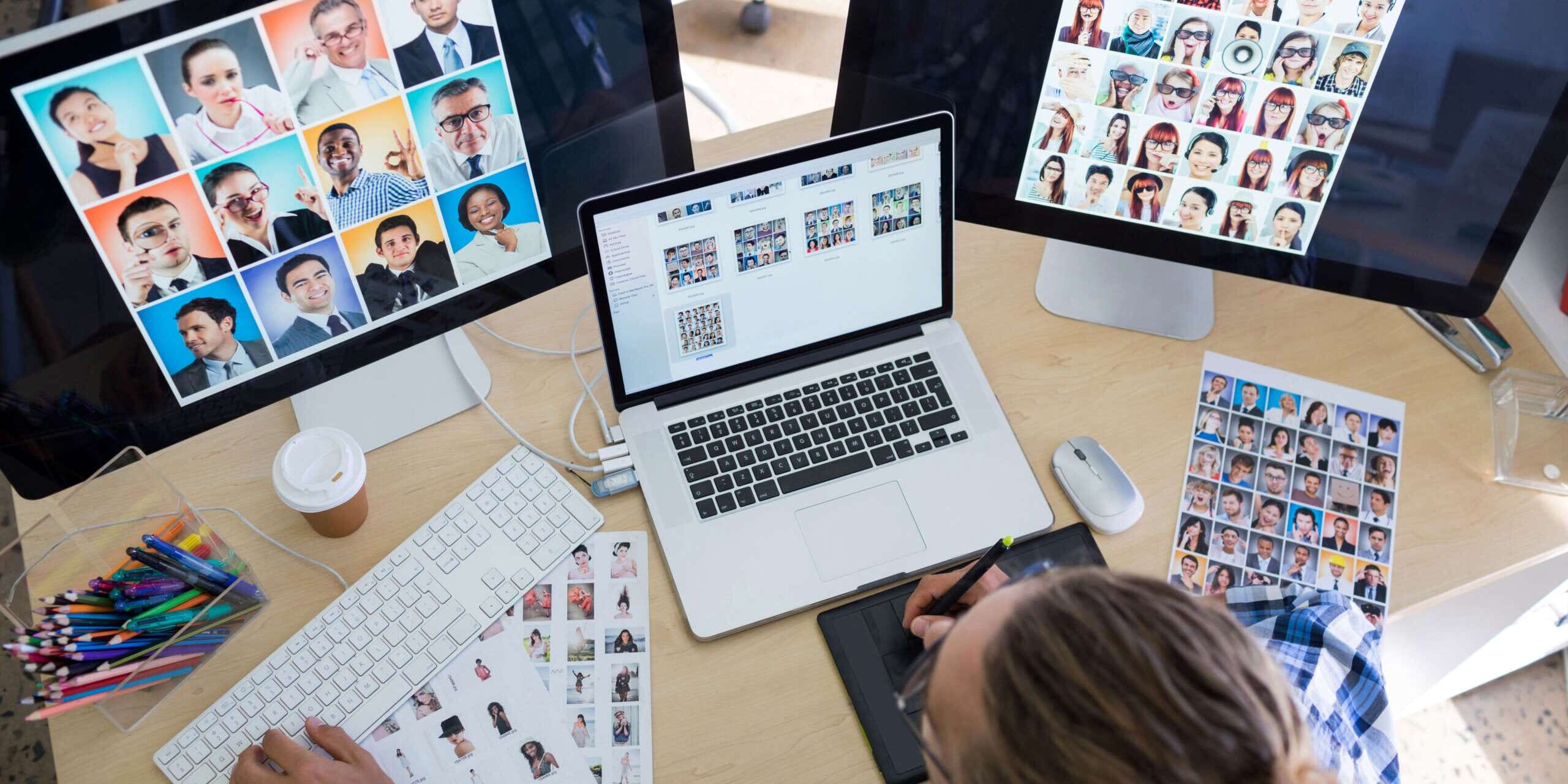 A person working at a desk with three monitors displaying multiple headshots. A keyboard and graphic tablet are on the desk with printed headshots scattered around. A cup of coffee and colorful pens are also visible.