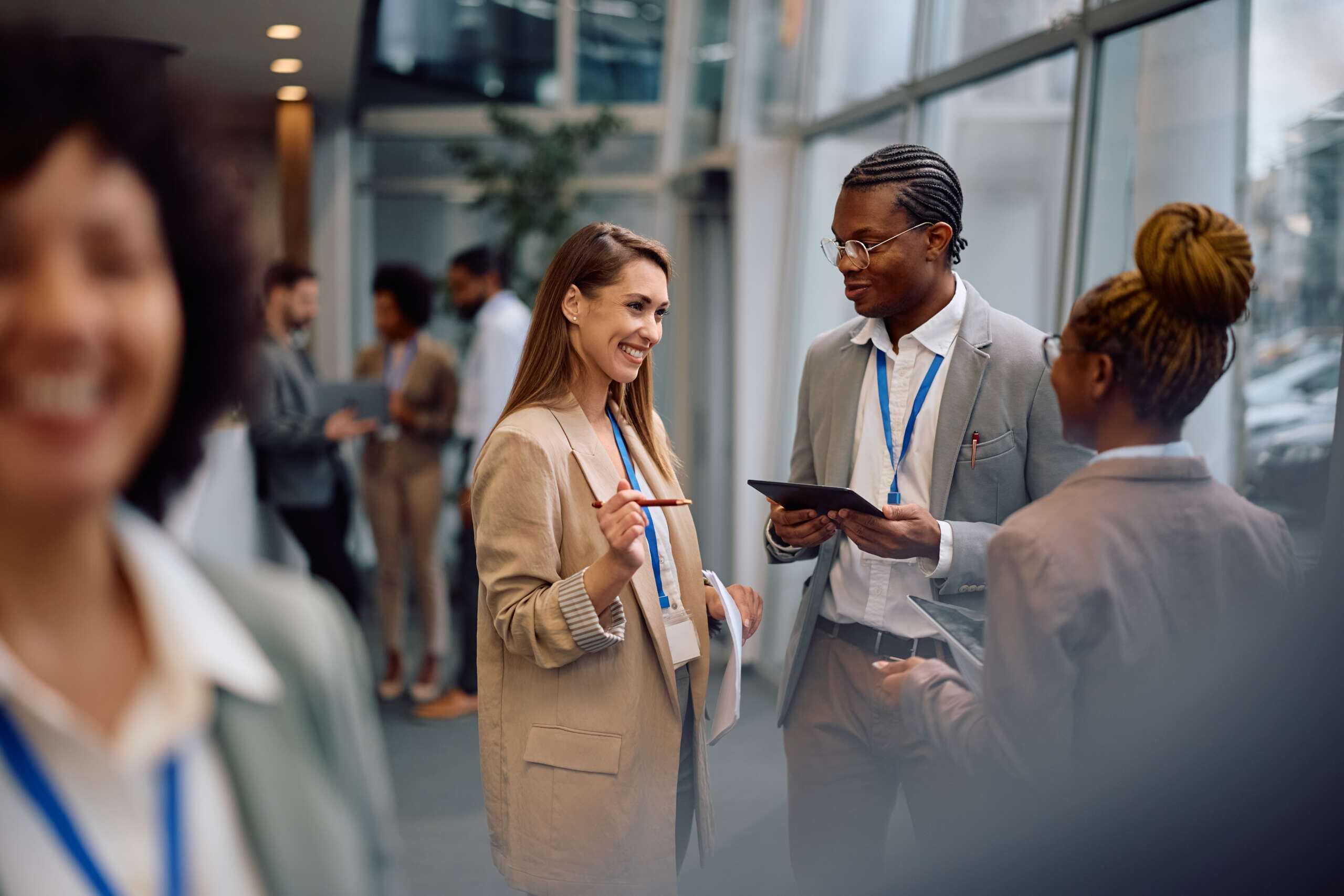 A diverse group of professionals in business attire engage in conversation at a networking event. They wear blue lanyards, and two people in the foreground hold a tablet and notebook while smiling. Blurred figures are visible in the background.