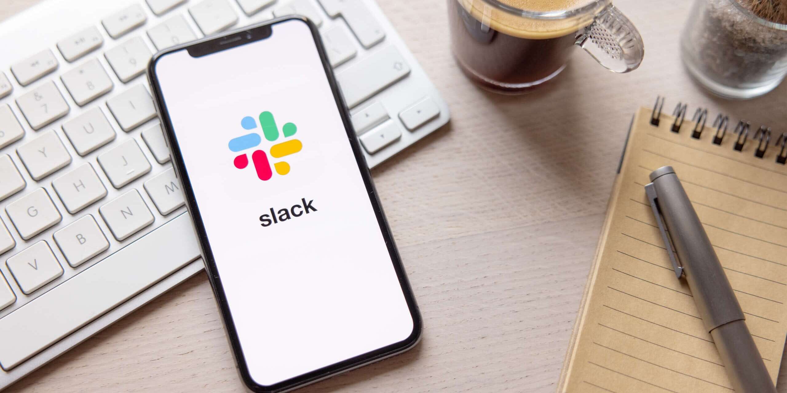 A smartphone displaying the Slack logo rests on a wooden desk next to a keyboard. To the right, there is a spiral notebook with a pen on top and a cup of coffee, suggesting a work environment.