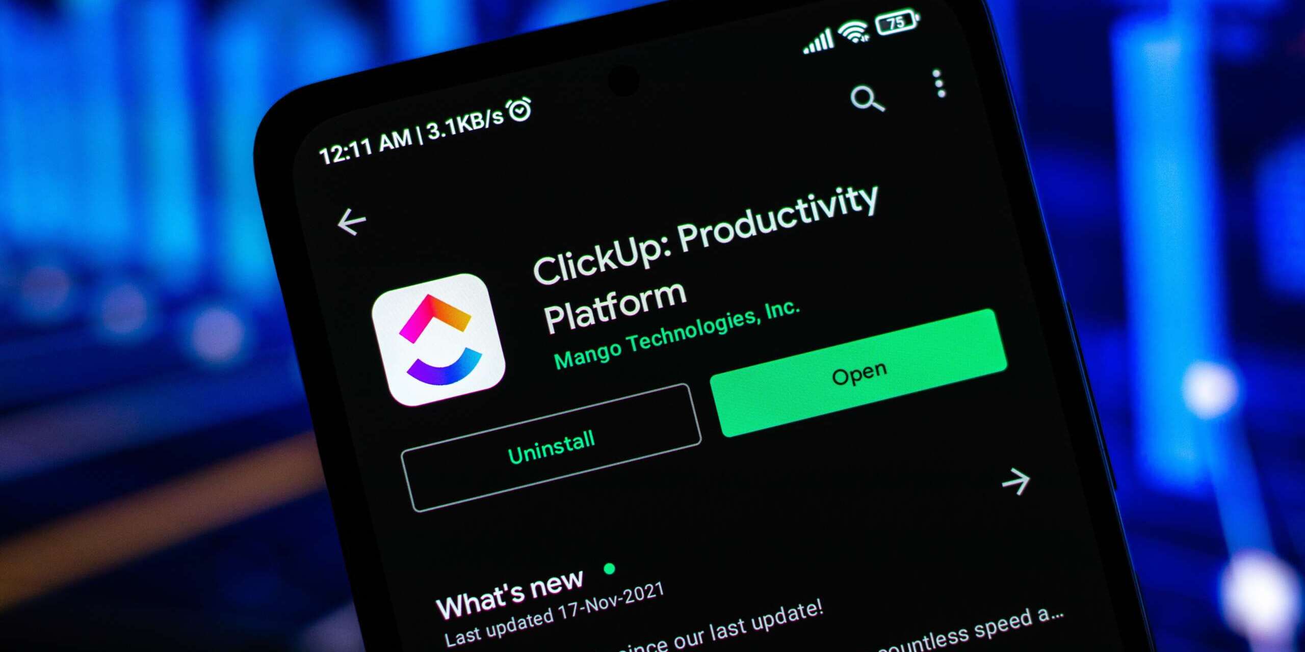 A smartphone screen displaying the ClickUp: Productivity Platform app on the Google Play Store. The apps icon and options to uninstall or open are visible. The background is blurred with blue lights.