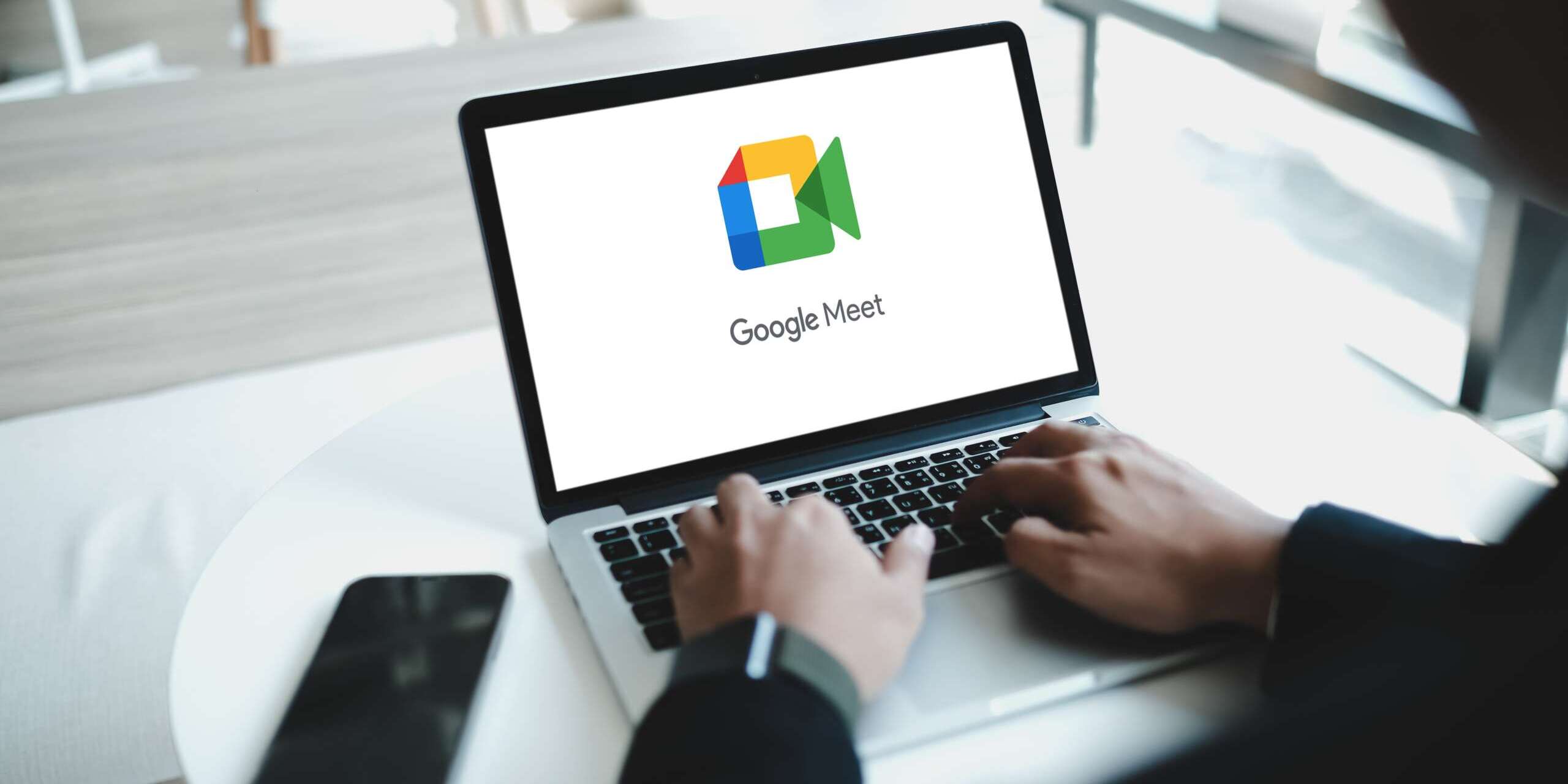 A person types on a laptop displaying the Google Meet logo. The laptop is on a round white table, with a smartphone placed nearby. The setting appears to be a modern office or workspace with large windows in the background.