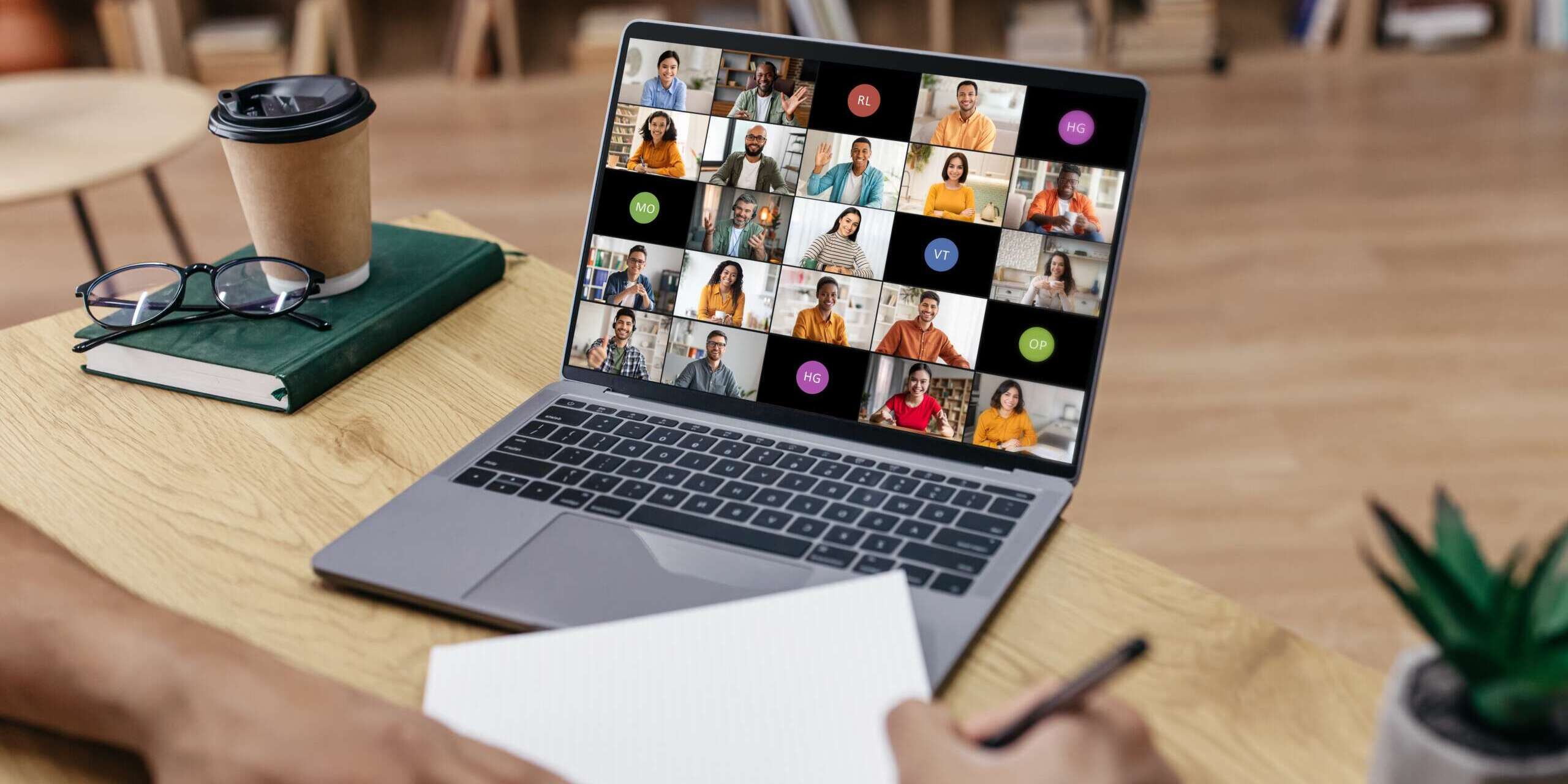 A person taking notes in front of a laptop displaying a virtual meeting with numerous participants. The setting is a well-lit room with a wooden desk, a coffee cup, notebooks, and a small plant. Shelves with books are in the blurred background.