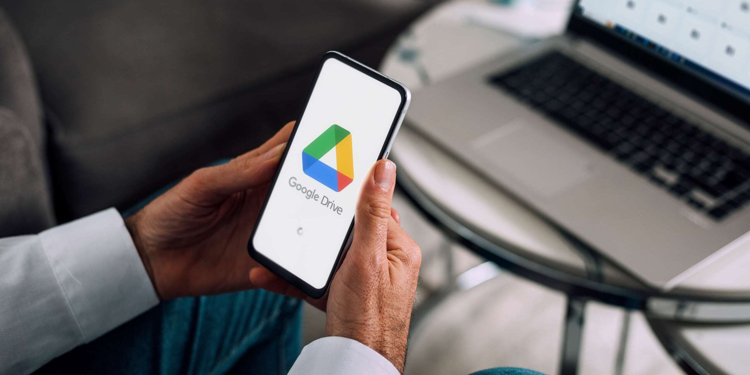 A person is holding a smartphone displaying the Google Drive logo. They are seated near a table with a laptop in the background. The setting appears to be a casual workspace or living room.