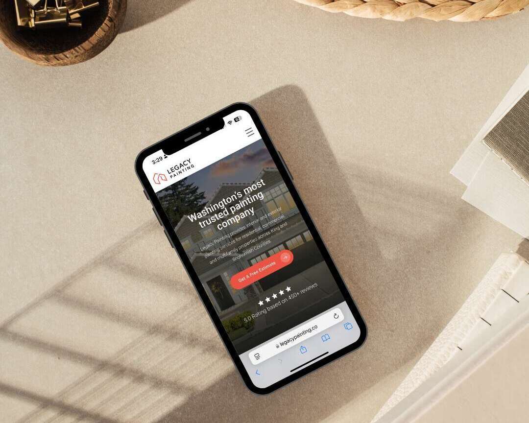A smartphone displaying a landscaping companys website lies on a beige surface near a woven basket, a glass container with binder clips, and an open notebook in sunlight.