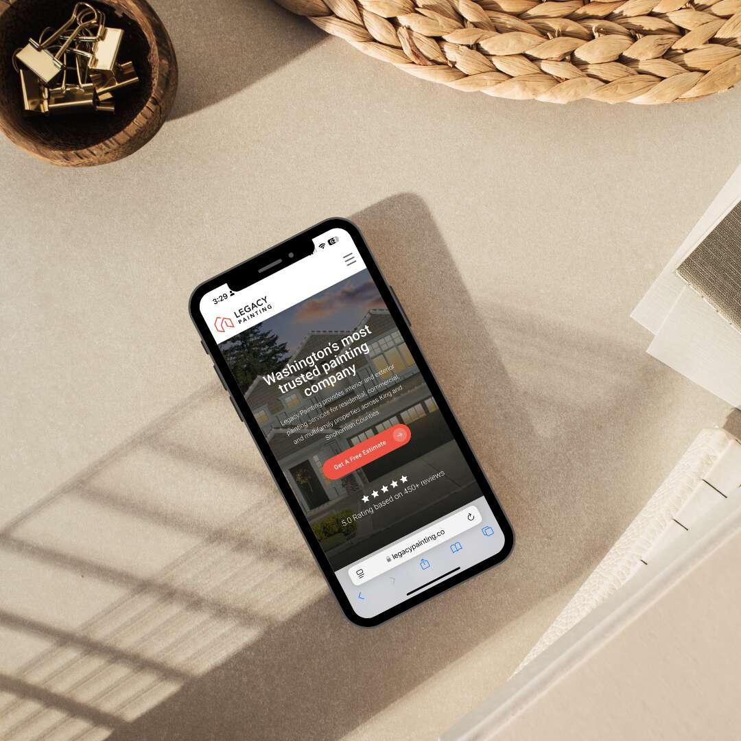 A smartphone displaying a landscaping companys website lies on a beige surface near a woven basket, a glass container with binder clips, and an open notebook in sunlight.