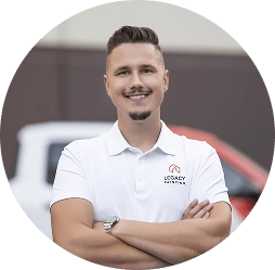 A smiling man with short hair and a goatee stands with arms crossed, wearing a white Legacy Painting polo shirt. The background is outdoors with blurred vehicles visible.