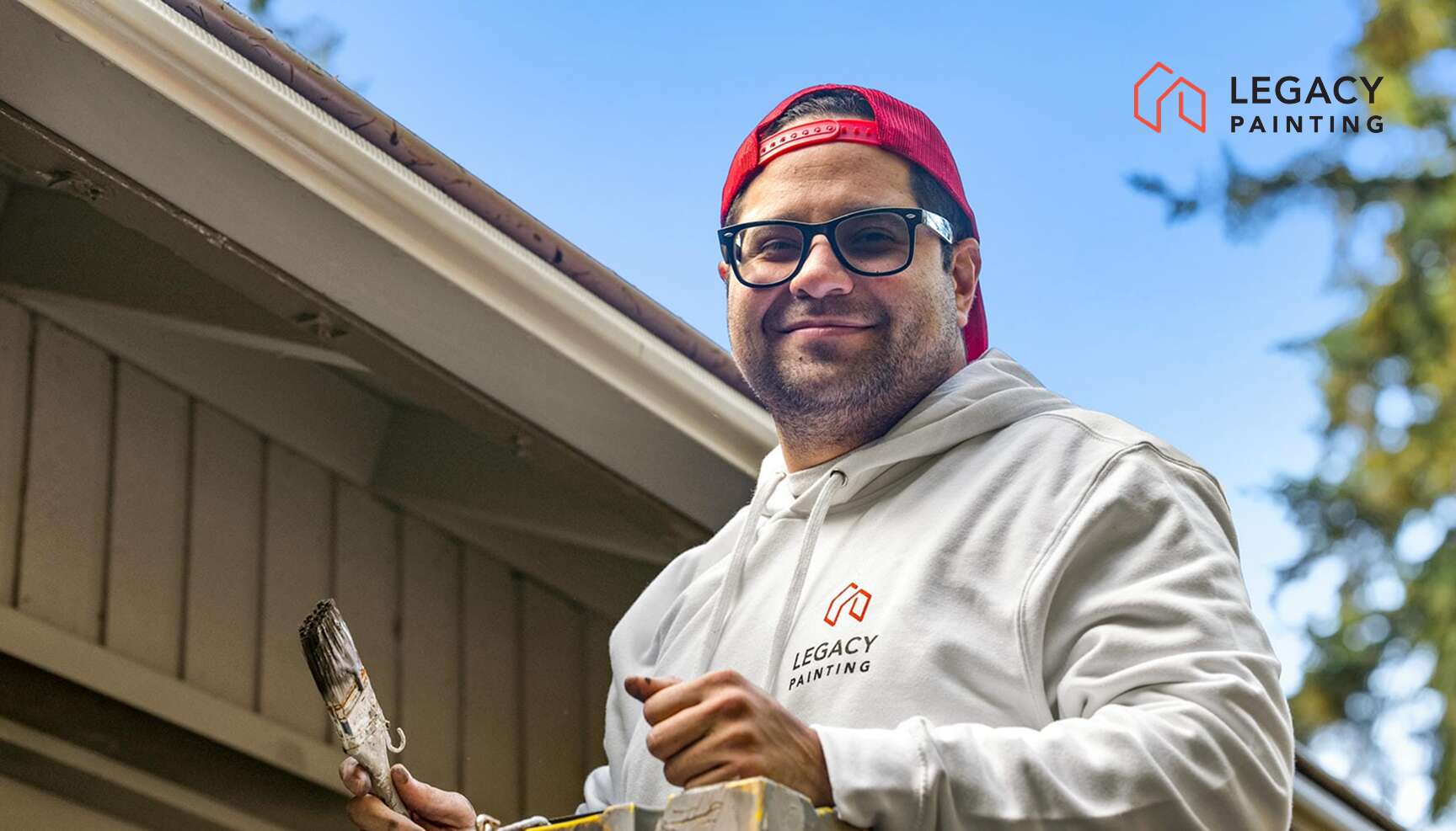 A man in glasses and a red cap, wearing a white Legacy Painting hoodie, holds a paintbrush and ladder outside a house. The Legacy Painting logo appears in the top right corner.