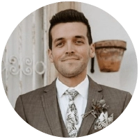 A man in a brown suit with a white shirt and patterned tie, smiling, stands in front of a rustic wall and a hanging flowerpot. He has short dark hair and is wearing a boutonniere.