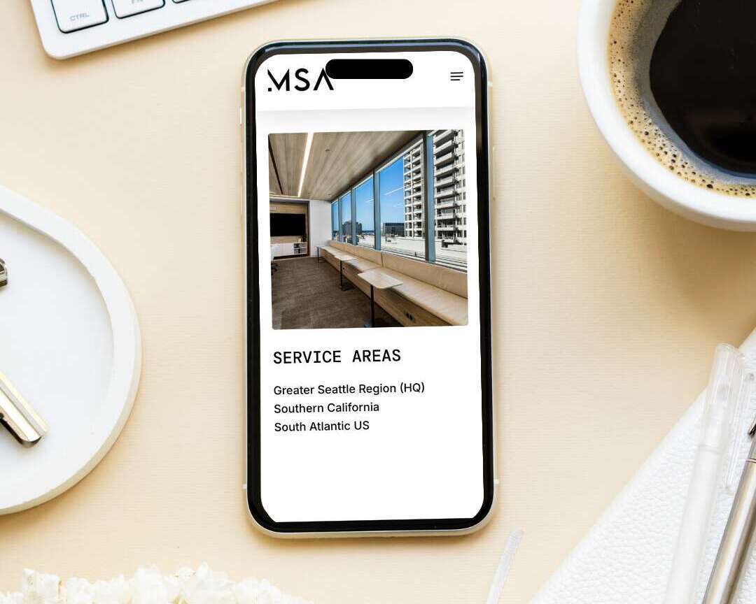 A smartphone on a desk displays a website featuring an office interior with a window view and a list of service areas. Surrounding items include a keyboard, coffee cup, pen, and flowers.