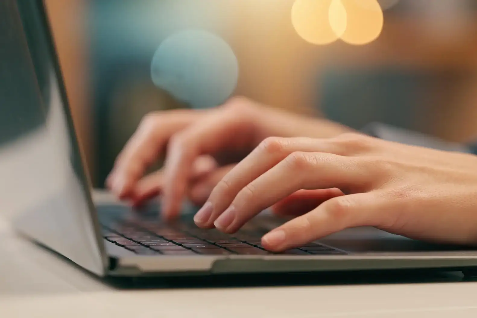 Close-up of a persons hands typing on a laptop keyboard, with a blurred background and warm lighting.