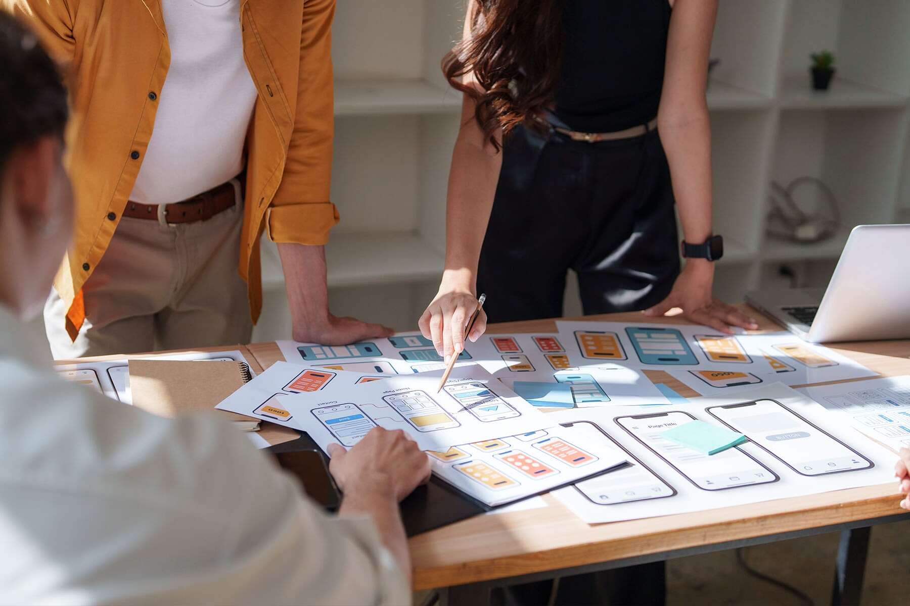 Three people standing around a table covered with paper prototypes and sketches of mobile app screens, discussing and pointing at the designs in a brightly lit office.