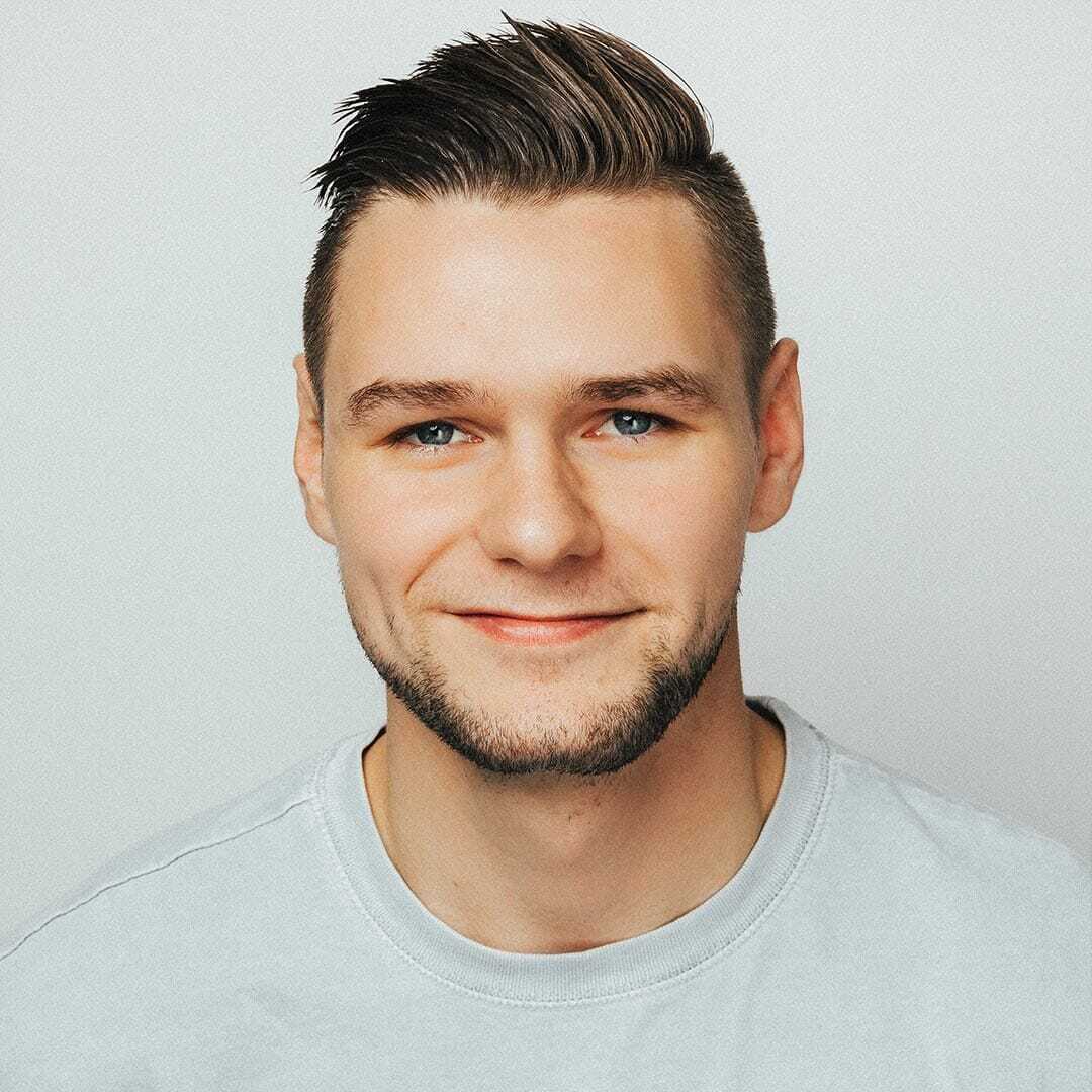 A young man with short, dark hair and a trimmed beard smiles at the camera. He is wearing a light gray T-shirt against a plain white background.