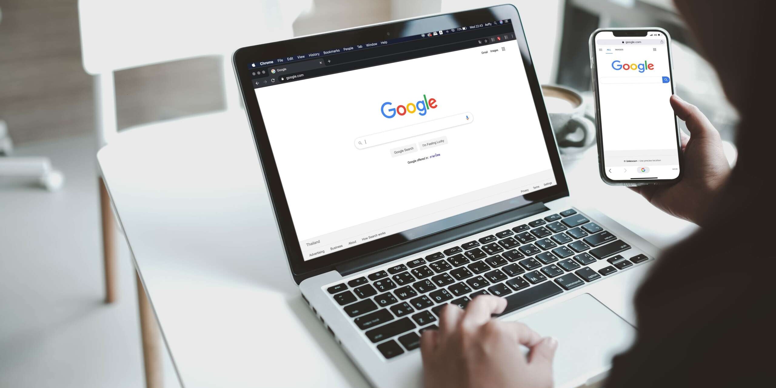 A person is using a laptop and holding a smartphone, both displaying the Google search homepage. The devices are on a white desk in a well-lit room with a white chair nearby.