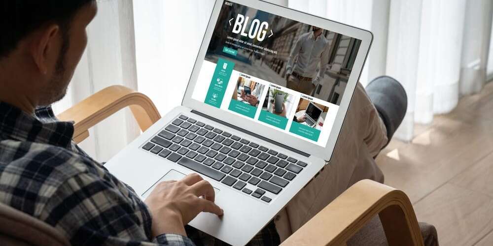 A person sitting in a chair, using a laptop displaying a website with the word BLOG at the top. The screen shows several images and text sections. The room has light curtains and a wooden floor. The person wears a checkered shirt.