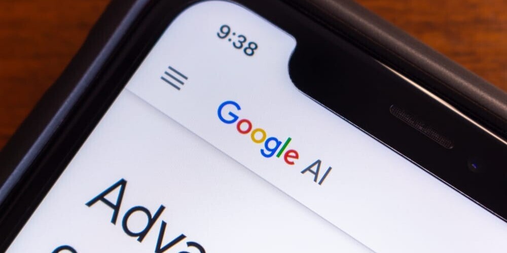 Close-up of a smartphone displaying the Google AI website. The screen shows part of the websites header with Google AI visible. The phones time reads 9:38, and it is set against a wooden background.