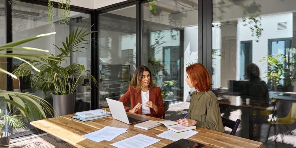 Two women are sitting at a wooden table in a modern, plant-filled office. They are discussing while looking at a laptop and documents. One woman is in a red blazer, the other in a green blouse. Large glass windows are in the background.